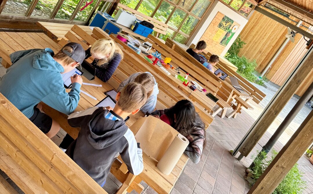 Auf einer Terrasse eines Holzpavillons sitzen Kinder an Holzbänken über Tablets gebeugt. 